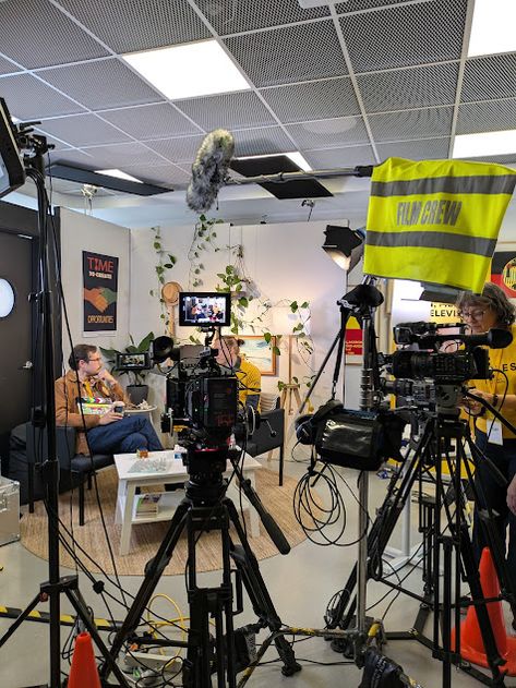 This contains an image of several people sitting in chairs around cameras and televisions on the floor, with one person holding a yellow flag