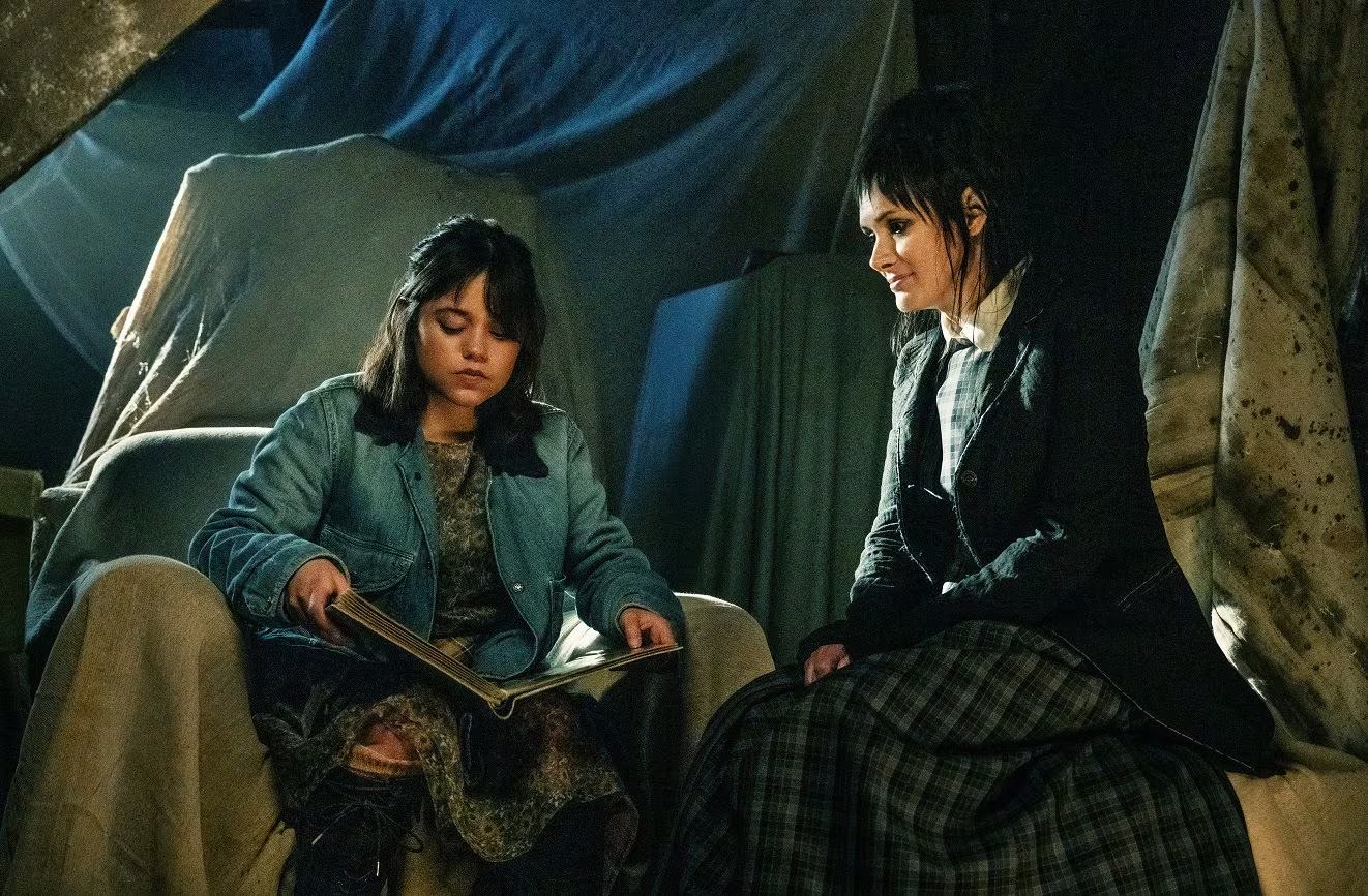 Jenna Ortega y Winona Ryder en una escena de Beetlejuice 2, sentadas juntas en un ambiente oscuro y misterioso.