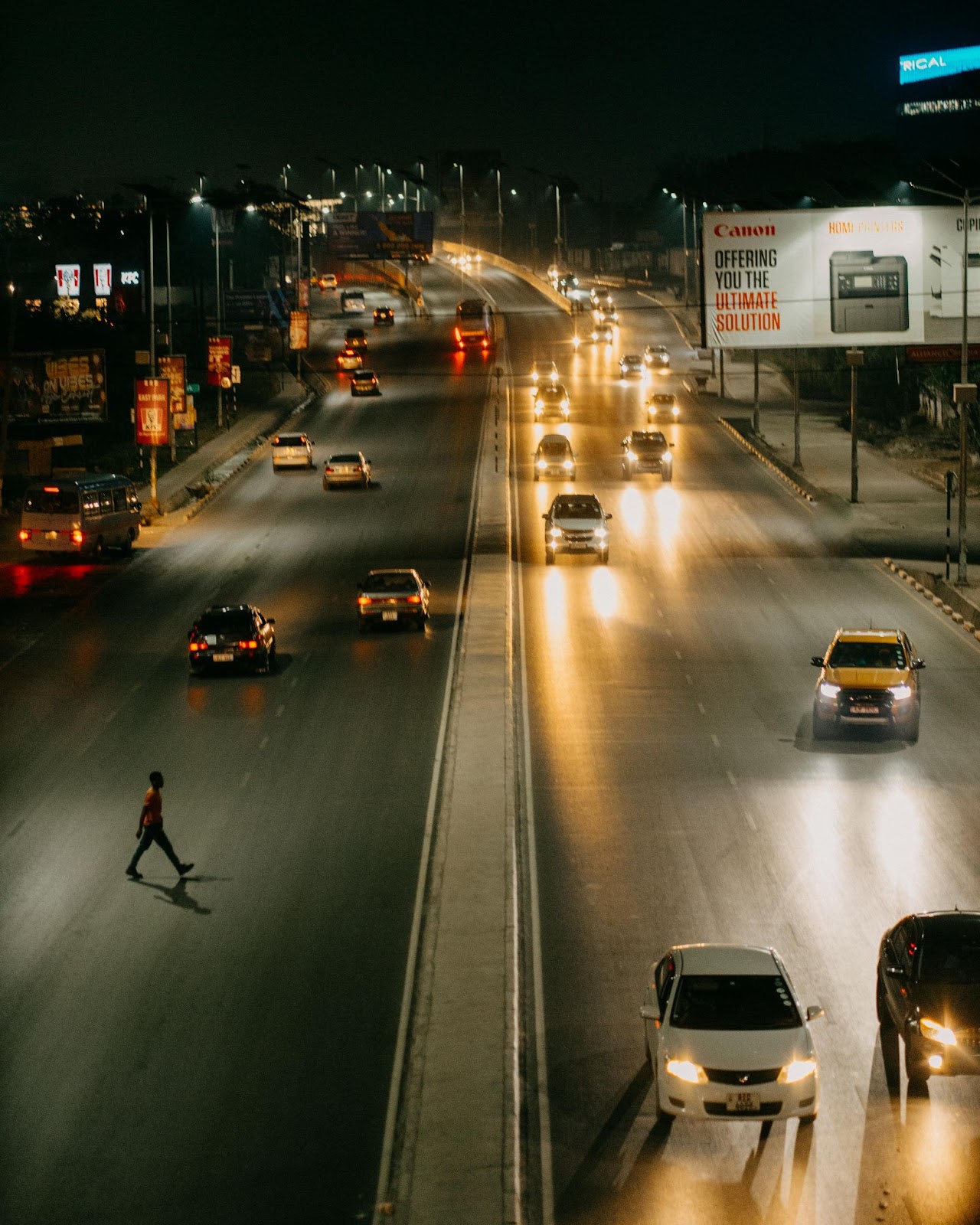 A night scene of a busy highway with cars driving on illuminated lanes and a pedestrian crossing the wide road, highlighting the risks of crossing streets in low visibility.