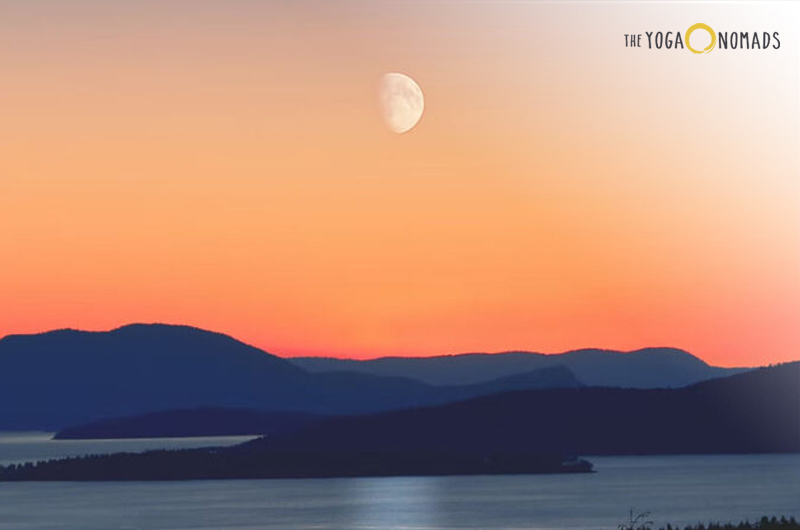 The image captures a scenic view of a sunset or sunrise over a body of water with silhouetted landforms, possibly hills or islands, in the foreground. The sky transitions from warm yellow and orange hues near the horizon to a deeper blue at the top. A partially visible moon is seen in the sky above the landforms.