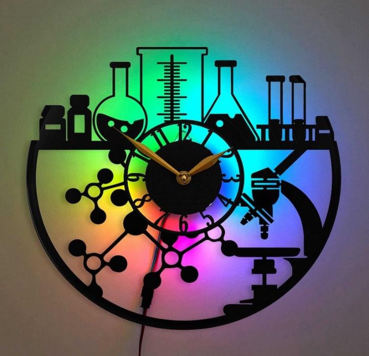 May include: A black and white clock with a rainbow background. The clock is shaped like a circle and has a black clock face with white numbers. The clock is surrounded by a black silhouette of a microscope, beakers, and molecules. The clock is a science-themed wall clock.