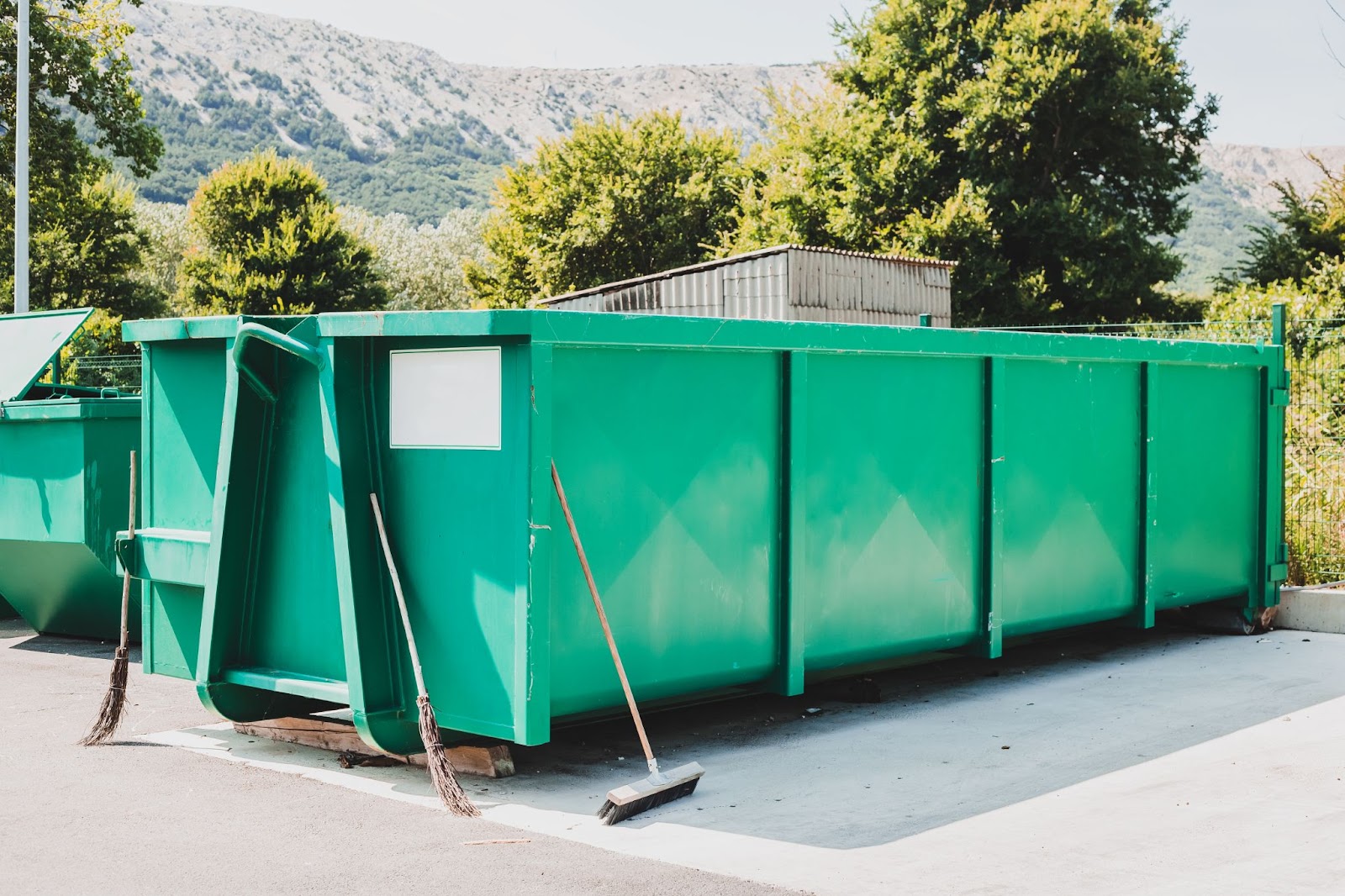 Dumpster outdoors next to brooms. 