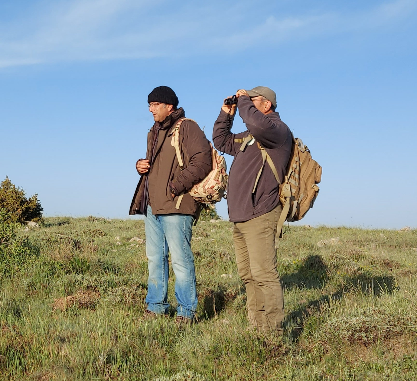 Two men standing in a field looking through binoculars

Description automatically generated