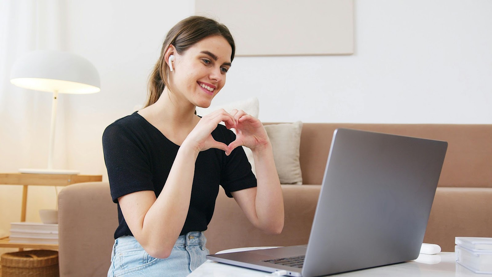 Long distance girlfriend sitting in front of the laptop, forming a heart with her hands and showing it to her boyfriend.