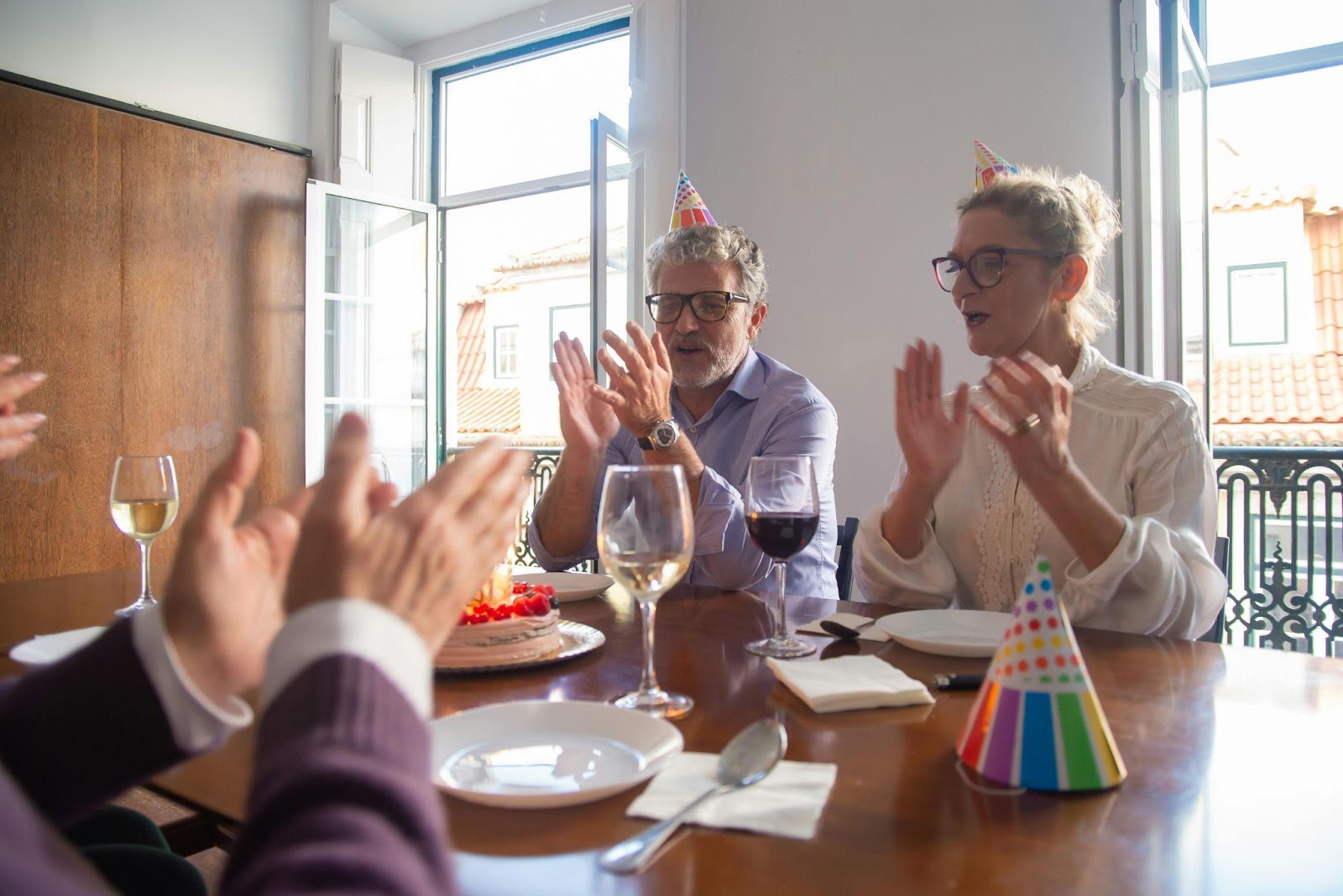 Older adults celebrating a birthday together at a sunny indoor dining table, wearing party hats, clapping, and enjoying each other's company with wine and cake.