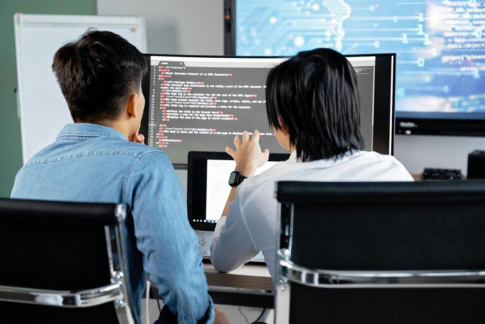 Two people sitting at a desk, focusing on a computer screen with lines of code.