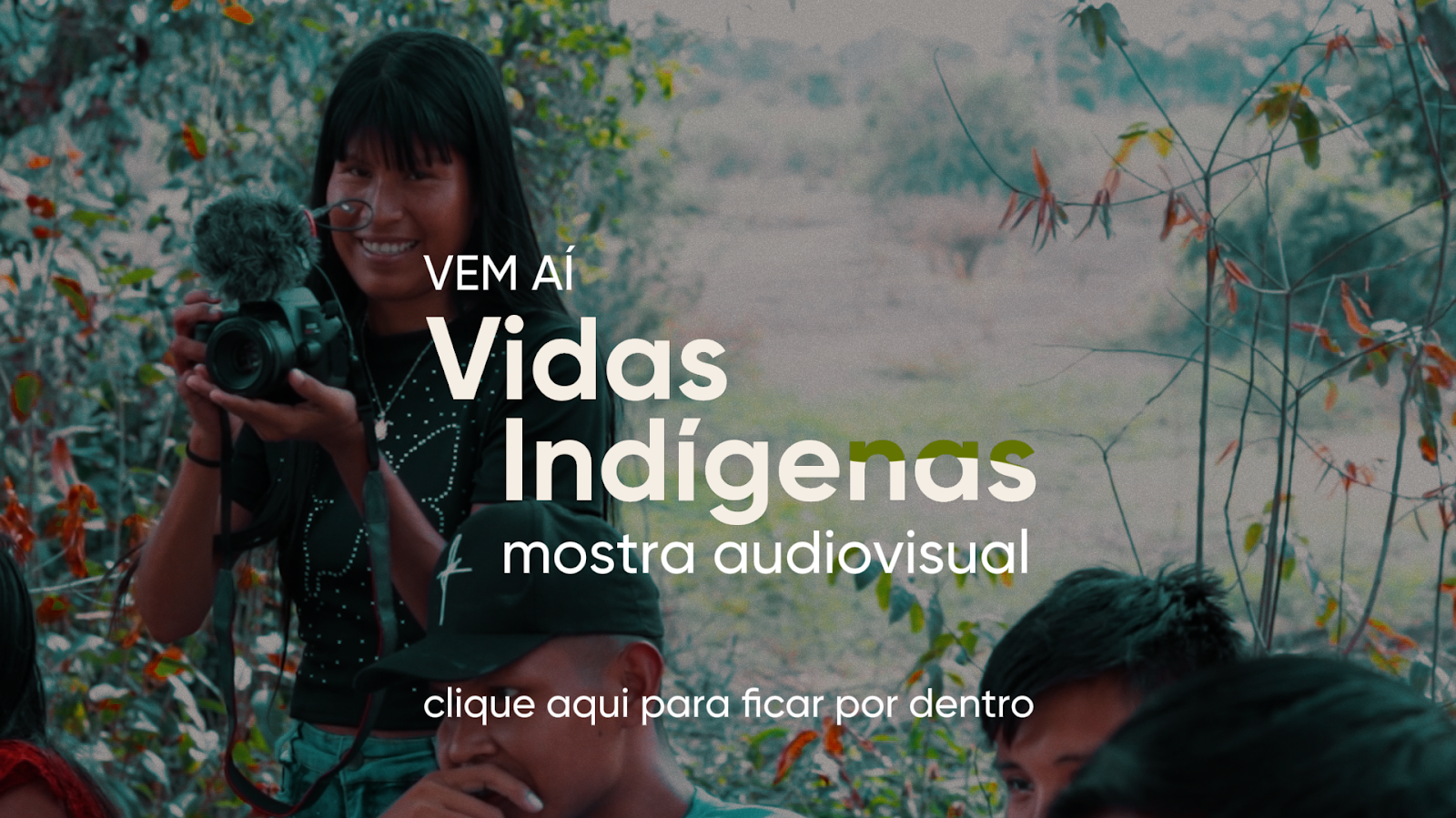Fotografia com filtro escuro aplicado. Nela, há uma moça indígena segurando uma câmera, dentro de uma mata. Está escrito: Vem aí Vidas Indígenas Mostra Audiovisual. Clique aqui para ficar por dentro.