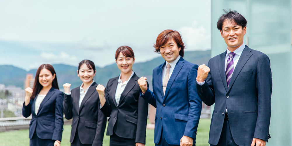 屋外でガッツポーズをとりながら並ぶスーツ姿の男女５名のスタッフ集合写真