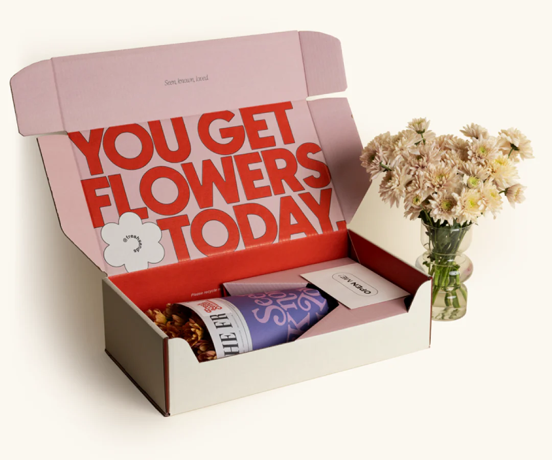 A Fresh Sends flower delivery box and an arrangement of white daisies in a cup of water.