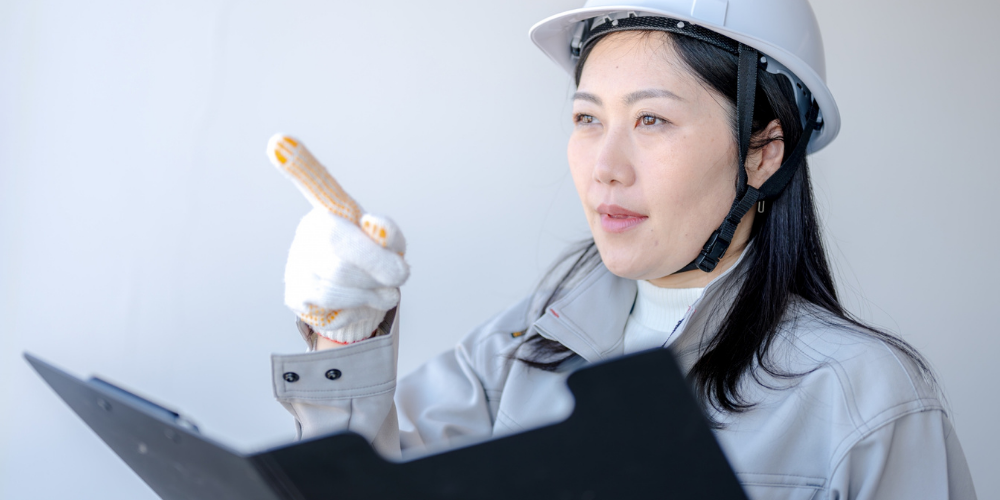  ヘルメット姿でクリップボードを持ち、指差しながら点検項目を確認する女性建築士