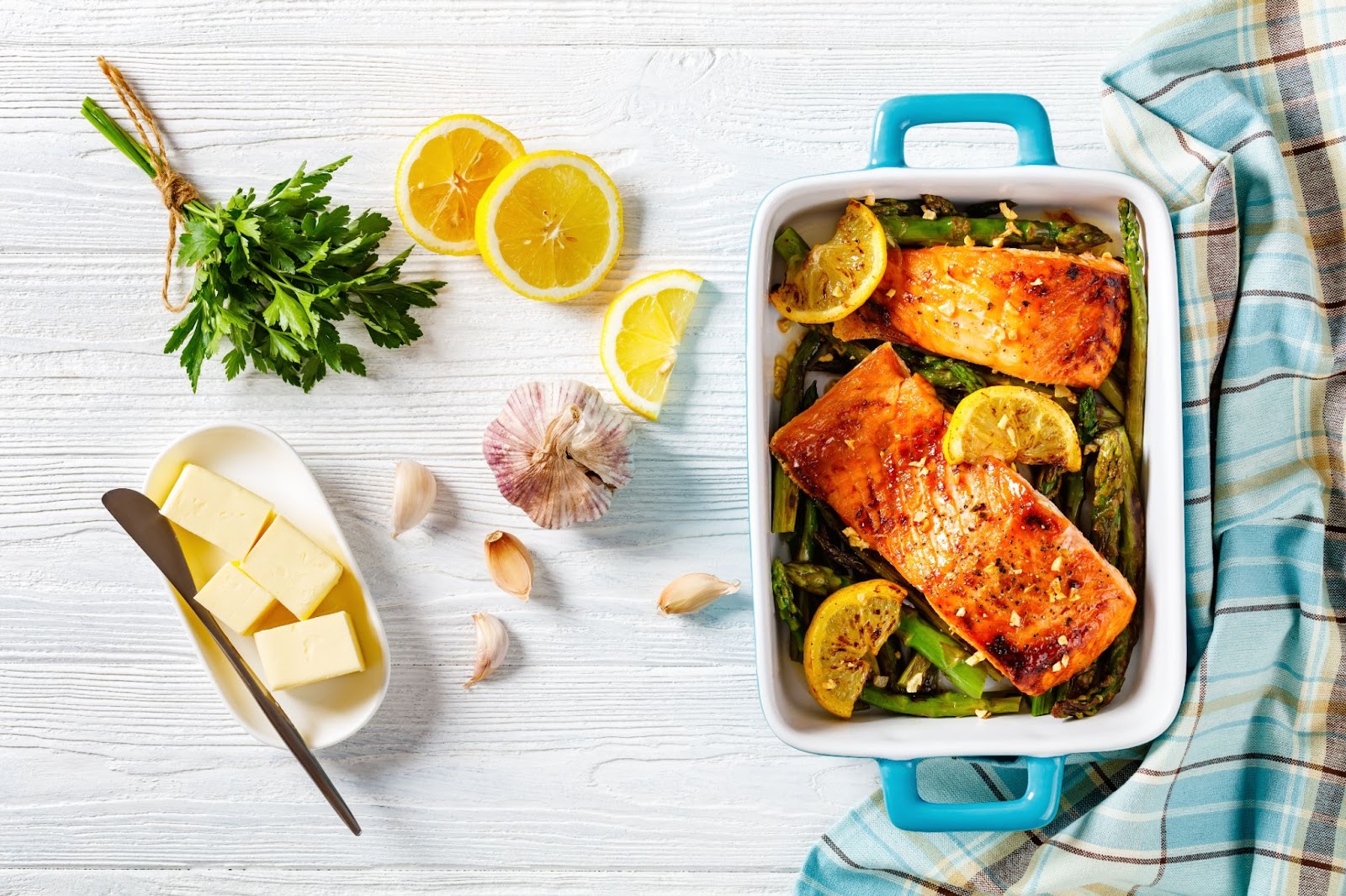 A pan of roasted salmon with asparagus and lemon placed next to ingredients: Parsley, lemon slices, garlic, and a tray of sliced butter.