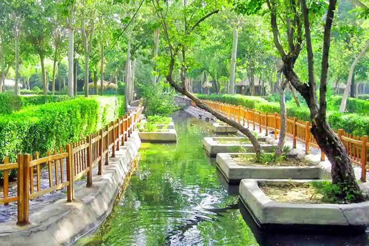 Scenic view of Shandiz, a popular tourist destination in Iran, featuring a tranquil waterway lined with lush greenery, wooden fences, and tall trees in a peaceful park setting.
