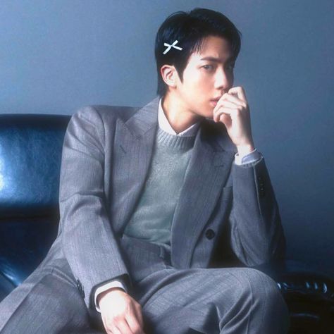 This  contain an image of Jin sitting on top of a blue couch wearing a suit and tie with a cross pin in his hair