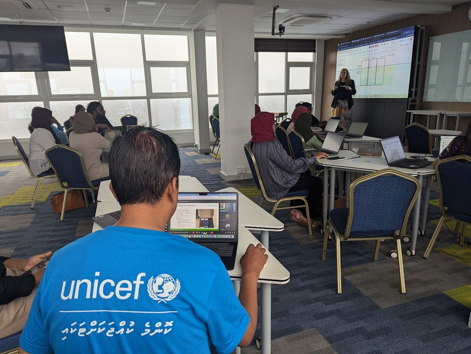 A classroom setting with attendees listening to a speaker.  A man with a UNICEF shirt sits in the foreground. 