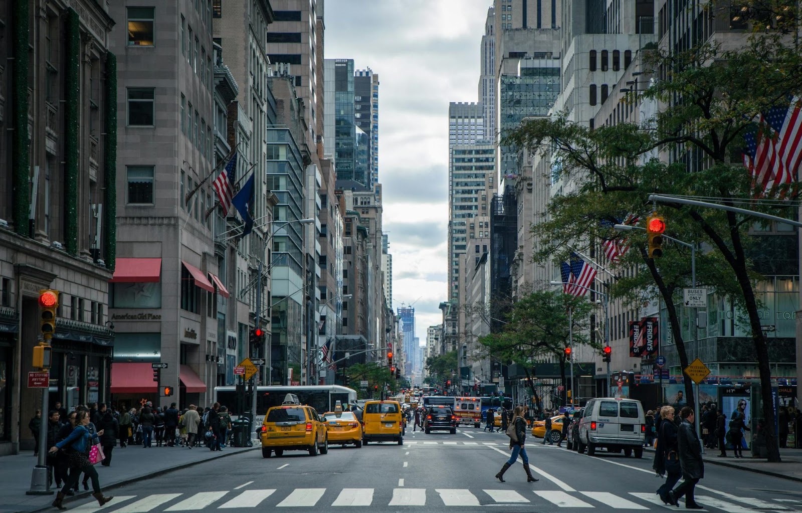 A bustling New York City street with tall buildings, yellow taxis, pedestrians crossing at a crosswalk, and flags lining the sidewalks.