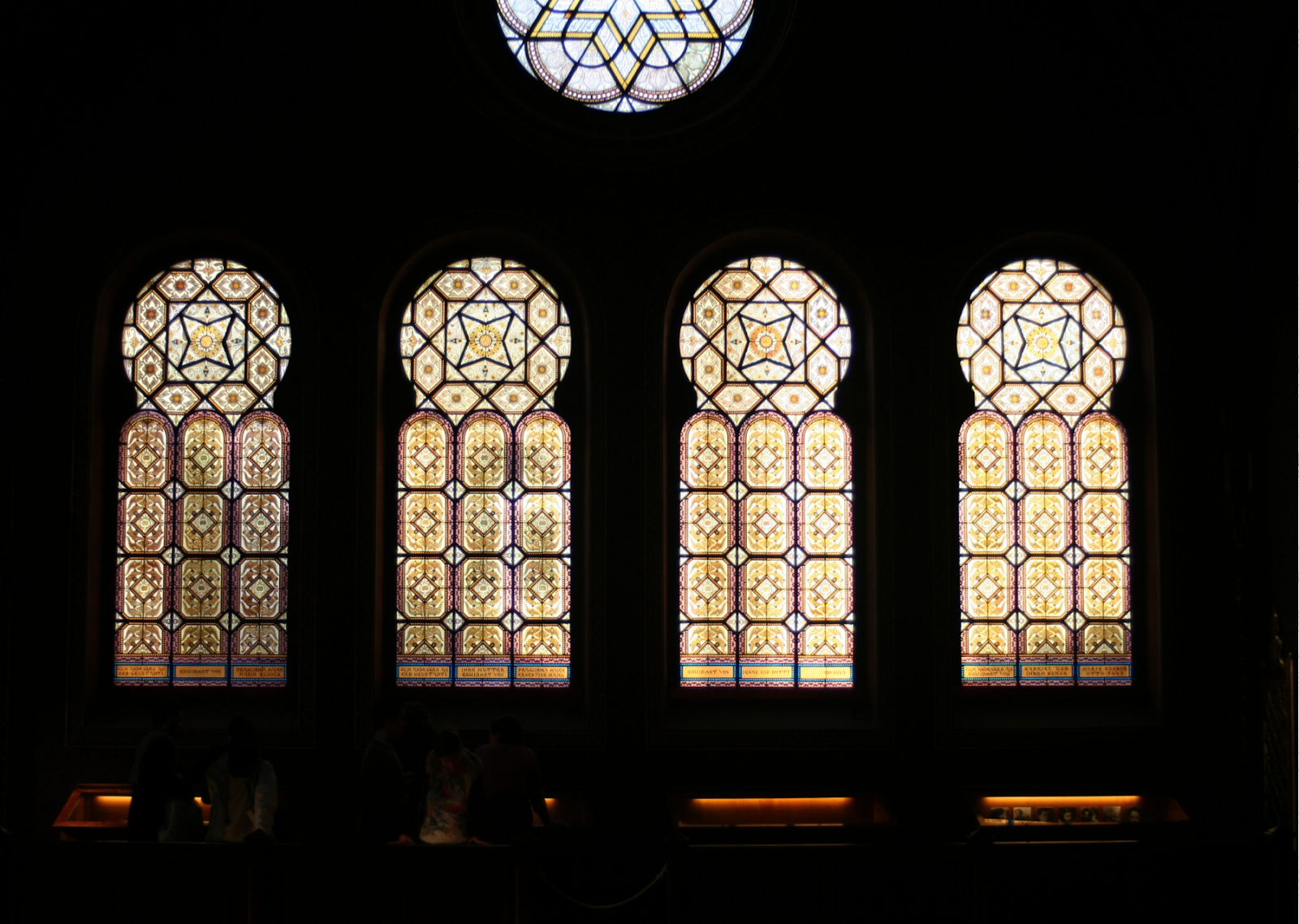 This image highlights an unspoken rule for tourists in Spain: showing reverence at sacred sites. It features four large, ornate stained glass windows with intricate geometric patterns and warm amber hues, casting a soft, golden glow in a dimly lit interior. Above, a circular stained glass window adds elegance with its complex design. Silhouetted figures at the bottom suggest a sense of scale and human presence, evoking quiet reflection and respect, essential when visiting Spain's historical and sacred locations.