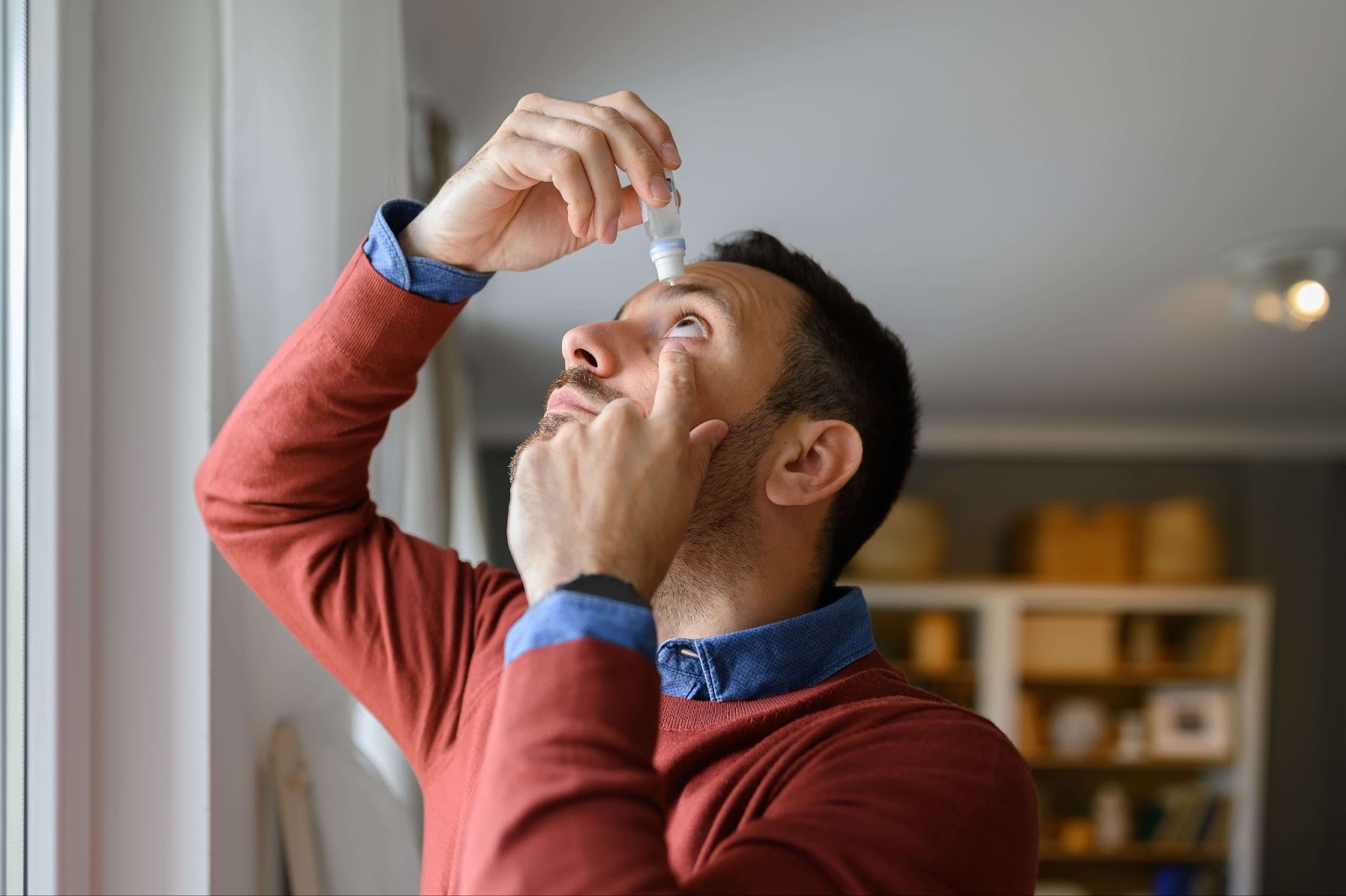 A person in a red sweater looking up while applying eye drops in a well-lit room.