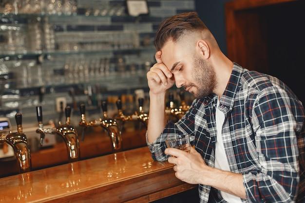 Man in a shirt holds a glass in his hands. Guy is sitting at the bar and holding his head.