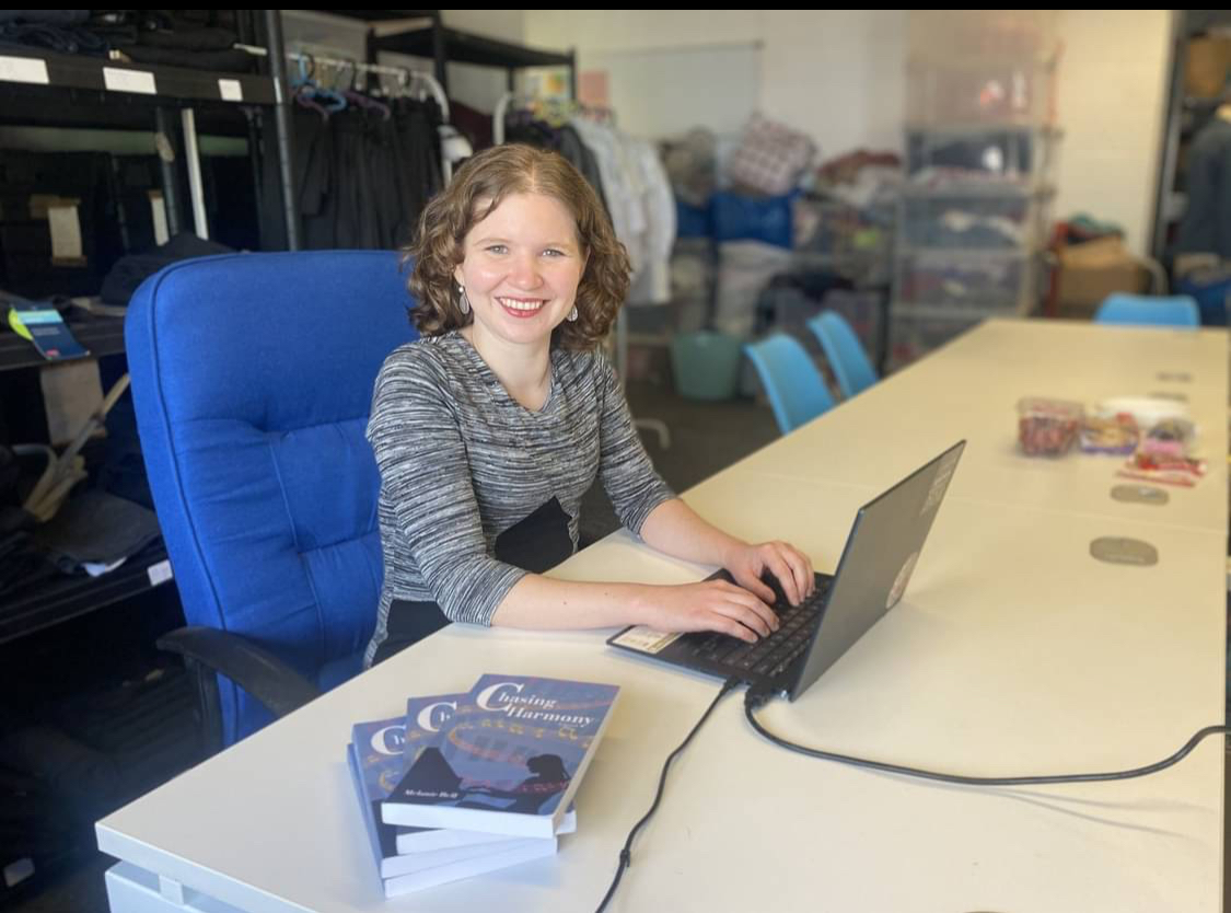 A woman smiles while working at a table with a computer. Office supplies and various items in the background. 