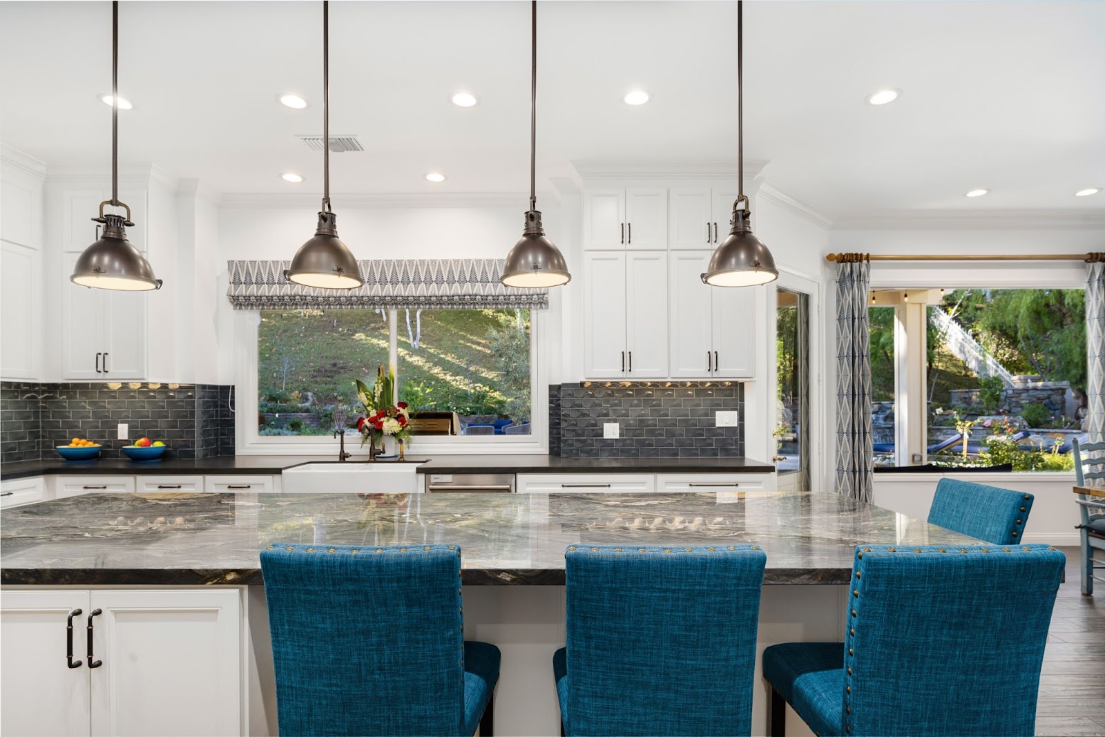 A brightly-lit kitchen featuring decorative backsplashes, a granite-topped counter, and a dining table with aquamarine cushioned seats, stainless steel lamps overhead, and wide glass windows overlooking the garden.