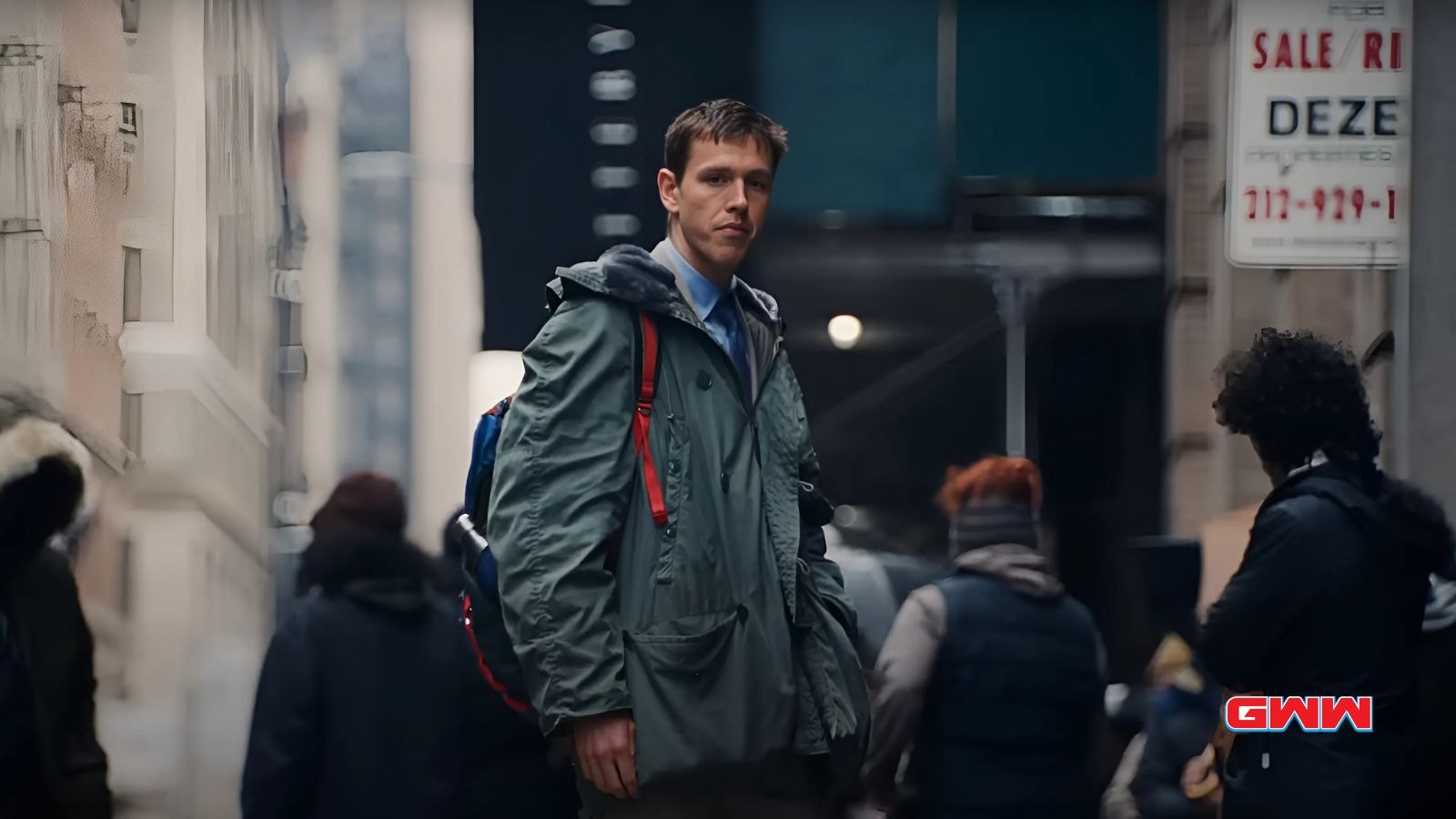 Harris Dickinson with short dark hair walking through a crowded, dimly lit urban environment.