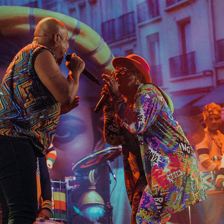Dois músicos brasileiros se apresentando juntos em um palco vibrante, vestindo roupas coloridas e alegres, enquanto cantam e tocam seus instrumentos durante uma apresentação musical ao vivo, celebrando a cultura musical brasileira.