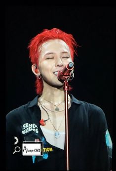 This  contain an image of  G-Dragon with red hair and piercings on his head singing into a microphone in front of a black background