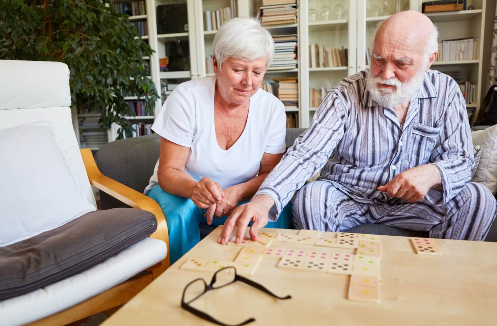 Older man and woman sitting on a couch and playing with dominoes on a coffee table.