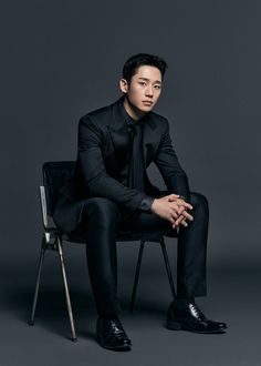 This contain an image of Jeon Hong Joon  in a black suit and tie sitting on a chair