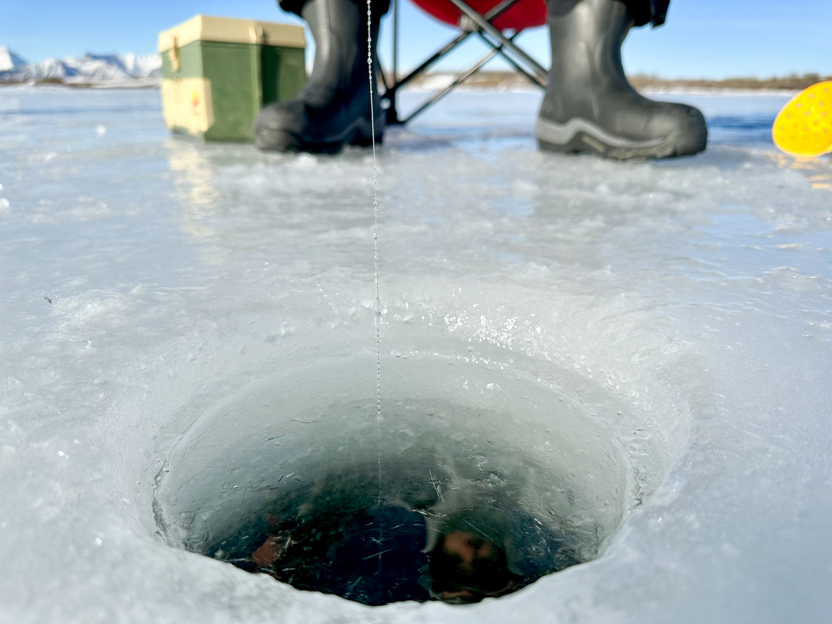 An ice fishing hole with black boots and a cooler behind.