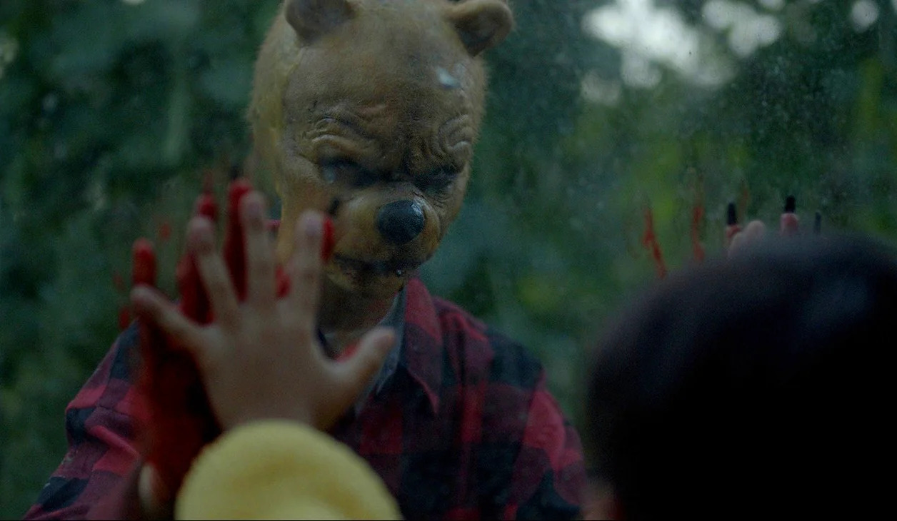 Persona con máscara aterradora de oso mirando a través de una ventana, mientras una mano ensangrentada se apoya en el cristal. Escena intensa y misteriosa en un ambiente de bosque.