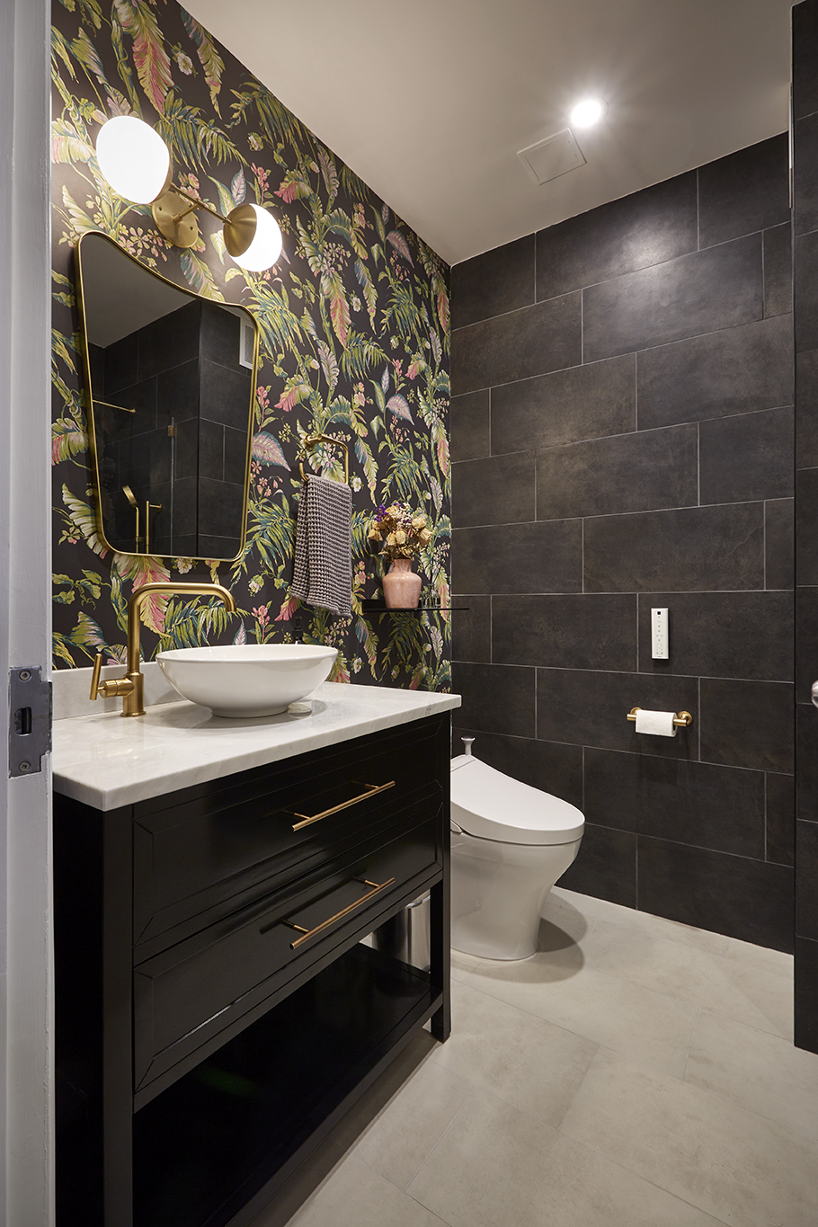 Powder room with floral wallpaper, black large format wall tile, beige floor tile, a large black vanity, and a vessel sink