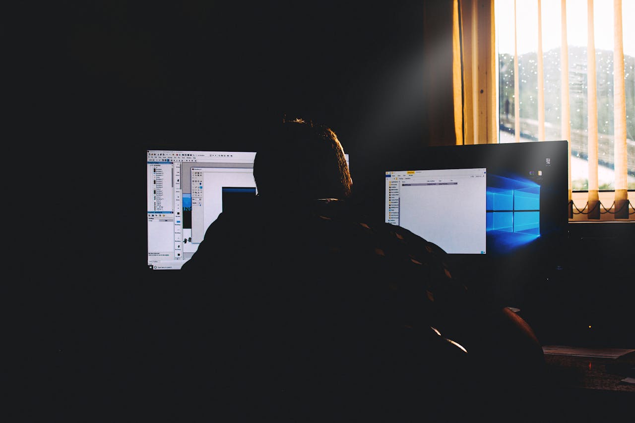 A person sitting at a desk working on multiple computer screens in a dimly lit room, illustrating how to protect computer systems from unauthorized access.