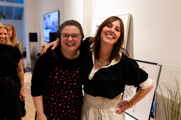 Cathlyn and Laura Belgray are standing close together indoors, smiling warmly at the camera. Cathlyn stands on the left has dark hair pulled back, wears glasses, and is dressed in a black top with a red and orange patterned dress. Laura stands on the right has long brown hair, is wearing a black blouse with rolled-up sleeves and a light-colored skirt, and has her arm around the Cathlyn. They are in a well-lit room with artwork leaning against the wall and a television screen in the background. Two other women are visible in the background, slightly out of focus, also appearing to enjoy the event.