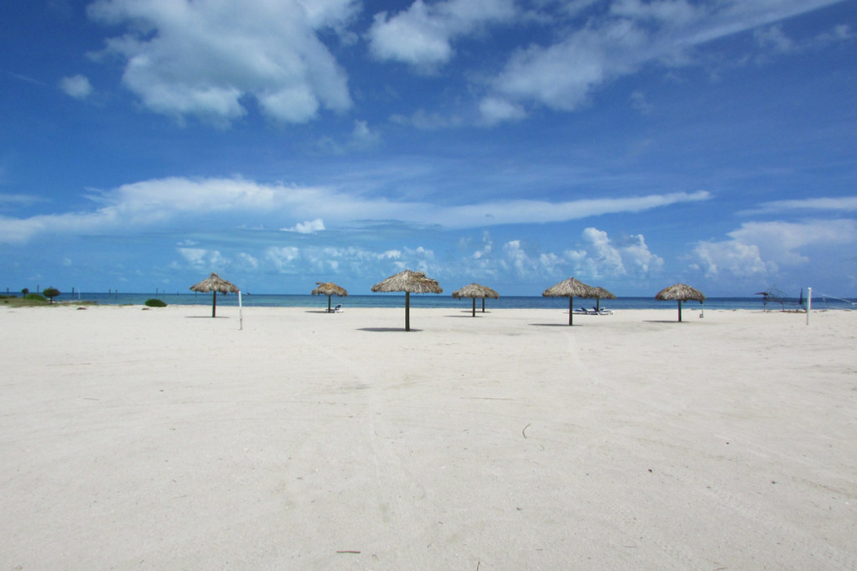 The beach features soft, white sand and is dotted with numerous umbrellas. The sky is clear and bright.