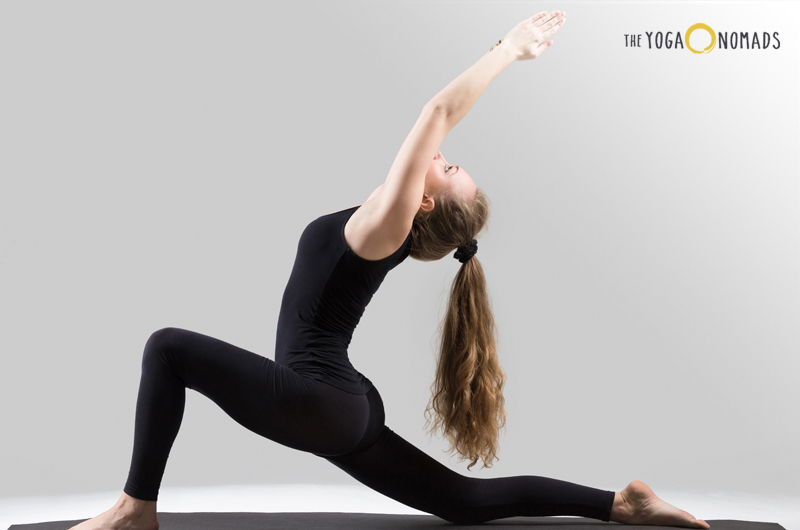 A person is performing a yoga pose on a yoga mat. The individual is in a lunge position with the right leg forward and the left leg extended back, both feet flat on the mat. The person’s arms are raised above the head, with palms facing each other and eyes looking upwards. The background is a plain, light grey color.