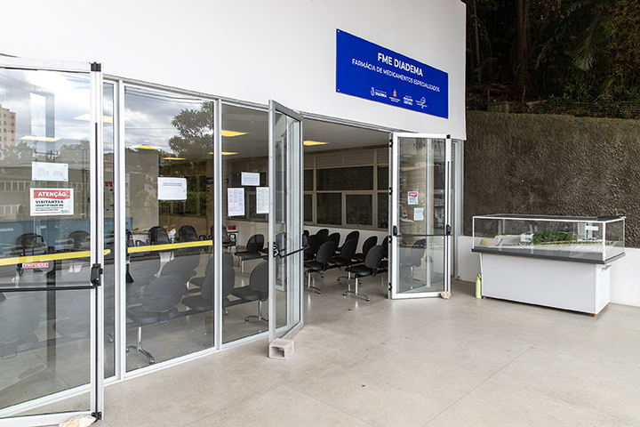 A imagem mostra a entrada de uma farmácia pública ou setor de medicamentos especializada, identificada pela placa azul no topo com o título "FME DIADEMA - FARMÁCIA DE MEDICAMENTOS ESPECIALIZADOS".  A entrada é composta por grandes portas de vidro transparentes, permitindo a visão do interior, onde há várias cadeiras pretas organizadas em fileiras, indicando uma área de espera para atendimento. Nas portas, há avisos e informativos impressos, incluindo um sinal de "ATENÇÃO" com informações direcionadas a visitantes.  Do lado direito da imagem, há um balcão ou vitrine com uma superfície transparente, que parece ser usado para exibição ou organização de algum material, provavelmente relacionado à farmácia.  O local é amplo, com piso claro e paredes brancas, proporcionando um ambiente limpo e organizado. Ao fundo, do lado externo, há uma área com vegetação natural, o que sugere que a farmácia está localizada em uma região com elementos naturais próximos.