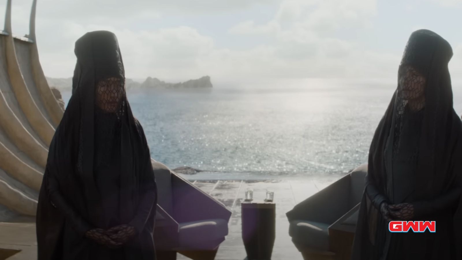 Two women in black veils, facing ocean, standing on terrace with chairs.