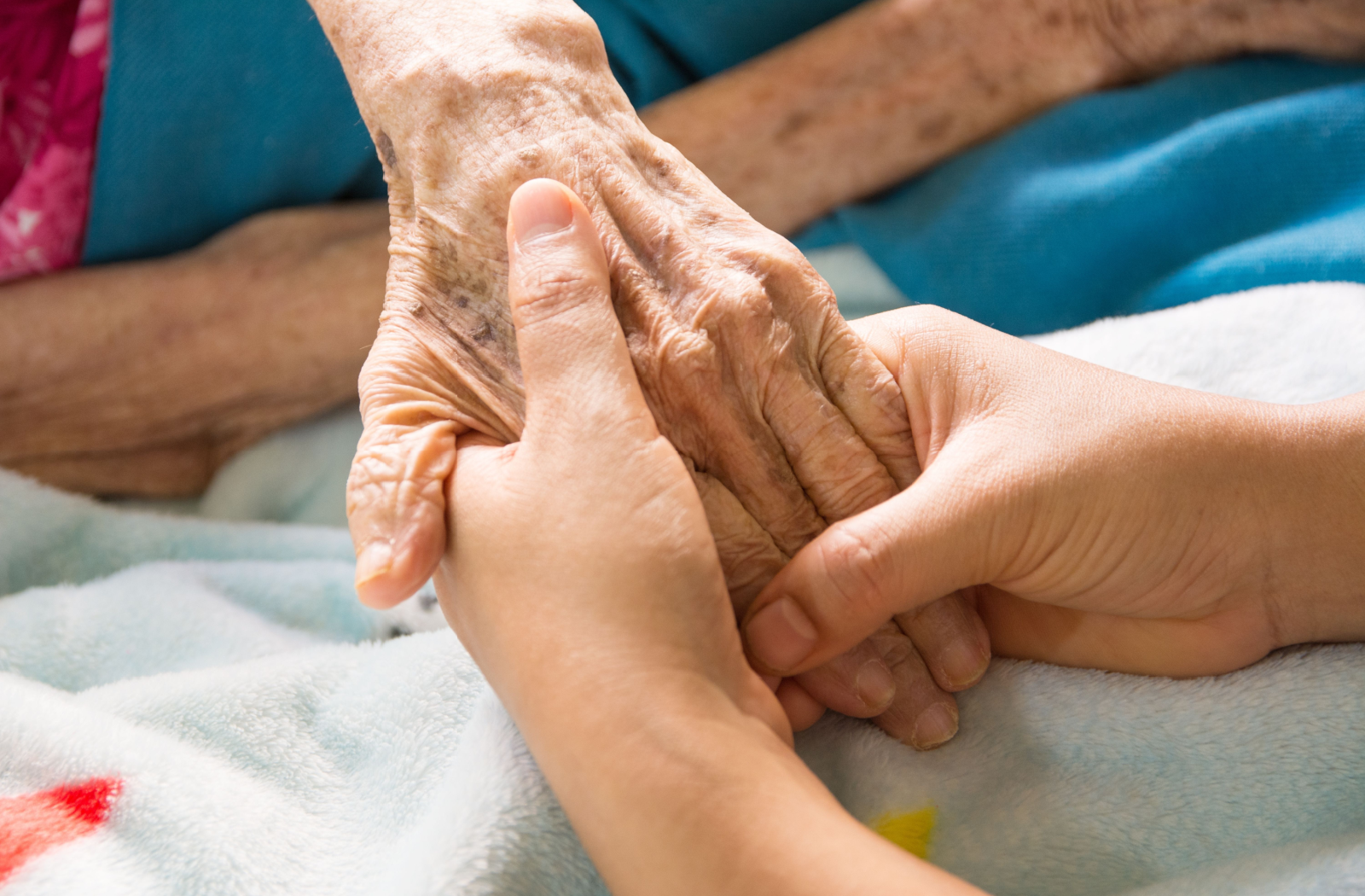 A senior and their adult child clasp hands over a cozy blanket at home.