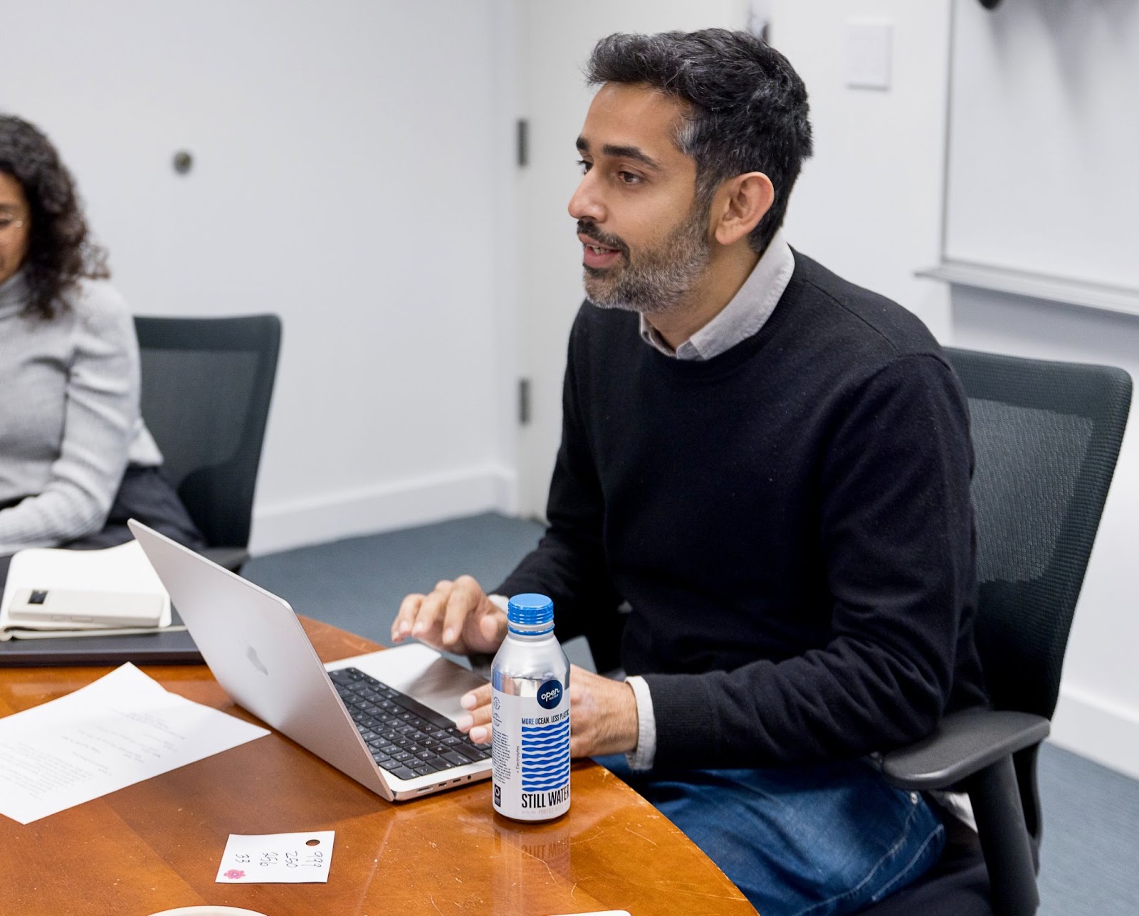 man sitting at a table with a laptop speaking to people out of frame