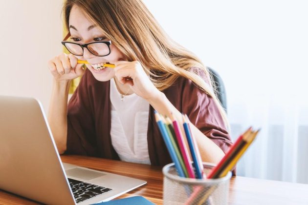 A woman with long hair and glasses is sitting at a desk, looking at a laptop screen with a stressed expression, while biting on a yellow pencil. A container with colorful pencils is on the desk.