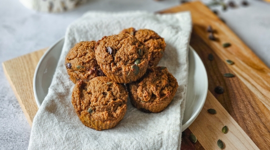 Une pile de muffins à la patate douce riches en fer sur une planche en bois, avec des graines de citrouille éparpillées et une tasse de café en arrière-plan.