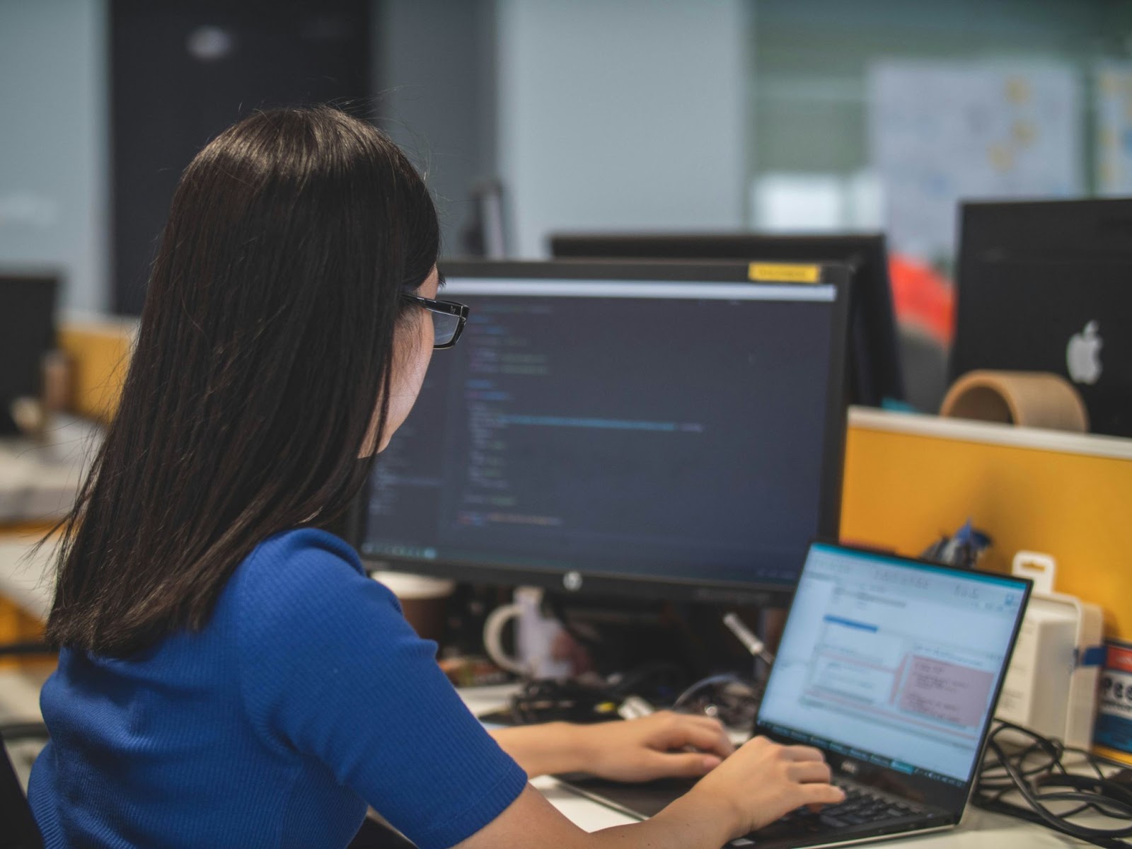 A woman in a blue shirt working on a laptop and desktop computer in an office.