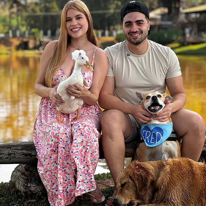 Casal de jovens sorrindo ao lado de cães e um pato em um parque, ambiente natural ao fundo, durante o dia ensolarado.