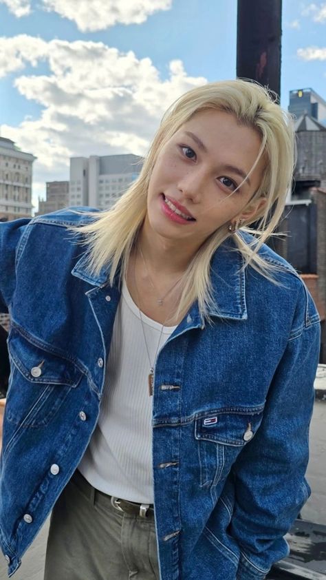 This  contain an image Stray Kids' Felix  with blonde hair wearing a denim jacket and khaki pants standing in front of a city skyline