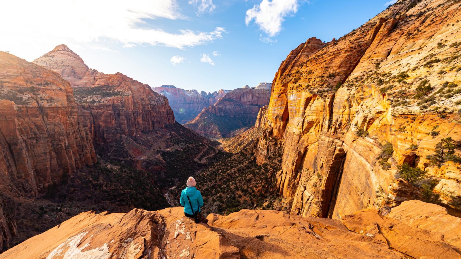 A girl in a blue jacket sits on a ledge overlooking a valley with sandstone cliffs on either side in Zion National Park