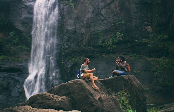 Two friends sit on rocks by a waterfall, one with a backpack, discussing taking a sabbatical leave to travel