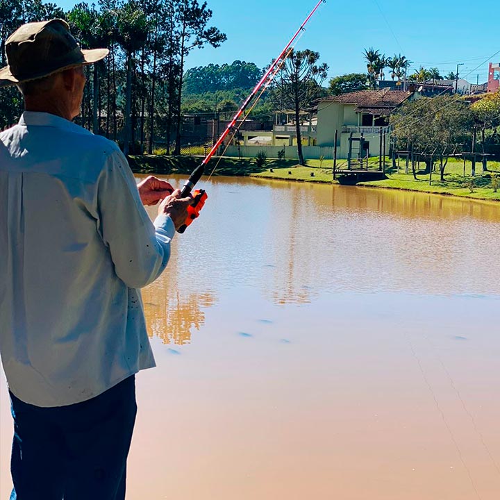 Pessoa pescando em um lago calmo com casas ao fundo e árvores, utilizando vara de pescar sob um céu claro, em um ambiente tranquilo e ao ar livre.
