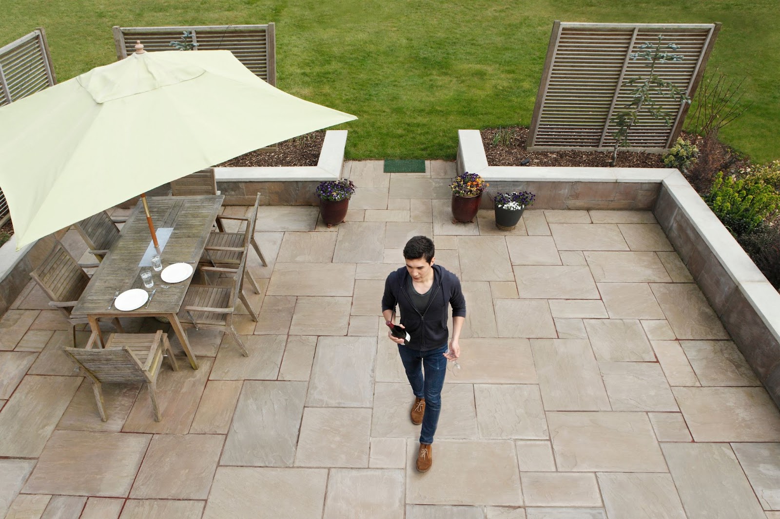 A man holding a wine bottle walks across a concrete patio with a pergola-covered dining table.