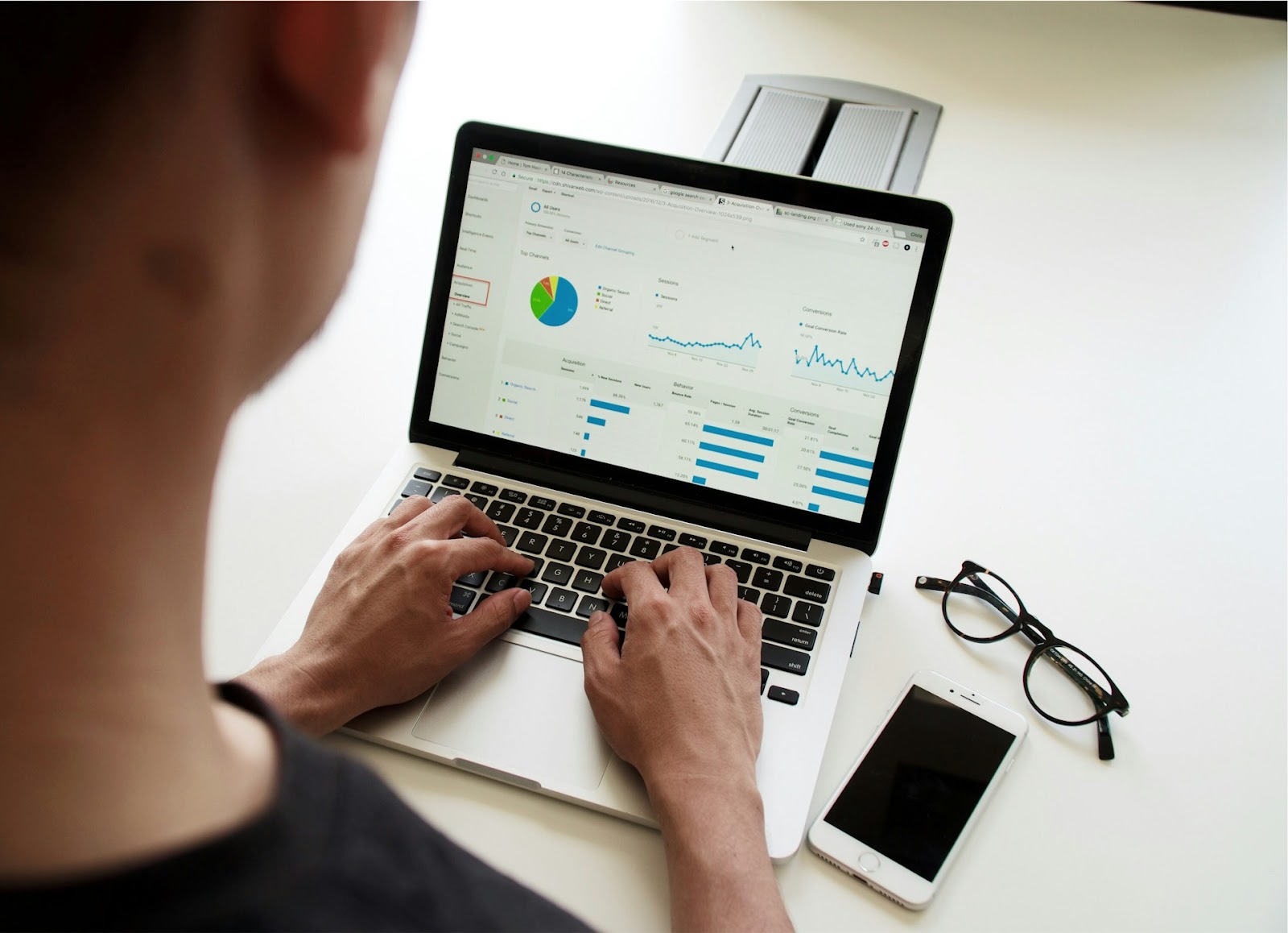 A person working on a laptop with analytical charts and graphs displayed on the screen, while a smartphone and glasses are placed nearby on the desk.